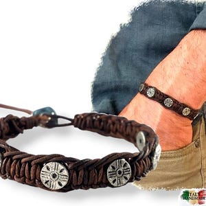 May include: A brown leather bracelet with silver metal accents. The bracelet is braided and features a decorative design on the metal accents. The bracelet is shown on a person's wrist.