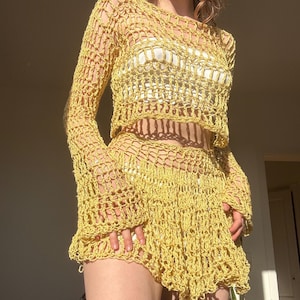 Könnte beinhalten: Gelbes Häkel-Top und -Shorts-Set. Das Top hat lange Ärmel und einen cropped Schnitt. Die Shorts haben einen Rüschensaum. Das offene Gewebe des Häkels erzeugt einen durchsichtigen Effekt. Das Set ist aus leichtem, sommerlichem Garn gefertigt.