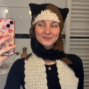 Könnte beinhalten: Gehäkeltes Katzenohren-Hut- und Schal-Set. Der Hut hat Ohren in Schwarz, Braun und Weiß. Der Schal ist cremefarben mit schwarzen und braunen Akzenten. Das Set ist aus weichem, strukturiertem Garn gefertigt. Der Schal ist etwa 150 cm lang.