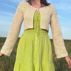 May include: A cream-colored, long-sleeved crochet bolero with a tie-front closure, worn over a lime green, button-down, sleeveless dress featuring a smocked bodice and a tiered skirt design that falls below the waist.
