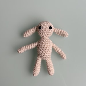 May include: A crocheted stuffed animal with a beige body and black eyes. The animal has long ears and a small, rounded body.