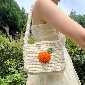 May include: A white crocheted shoulder bag with an orange crocheted fruit design. The bag has a long strap and is worn over the shoulder.