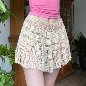 Könnte beinhalten: Cremefarbener Häkelrock mit ausgestellter Silhouette. Der Rock besteht aus einem locker gewebten Material, das ein offenes, luftiges Design schafft. Der Rock wird vor dem Hintergrund eines Holzschranks und grüner Pflanzen gezeigt.