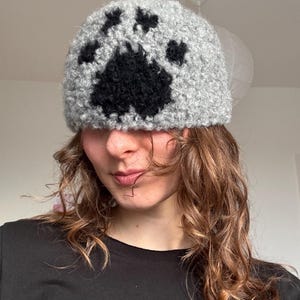 Könnte beinhalten: Graue gehäkelte Mütze mit einem großen schwarzen Pfotenabdruck-Design. Die Mütze besteht aus weichem, strukturiertem Garn. Diese Mütze ist zum Tragen auf dem Kopf konzipiert und ein gemütliches Accessoire.