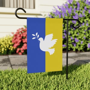 Puede incluir: Una bandera de jardín azul y amarilla con una paloma blanca que sostiene una rama de olivo. La bandera está unida a un poste de metal negro.