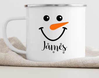 May include: White enamel mug with a snowman face and the name "James" printed on it. The snowman has black eyes, a black smile, and an orange nose.