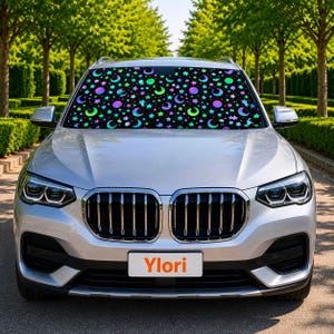Op de afbeelding: Een zilveren auto met een zwarte voorruitzonwering met een patroon van kleurrijke manen, sterren en cirkels. De zonwering is ontworpen om zonlicht te blokkeren. De auto heeft een witte nummerplaat met het woord "Ylori".