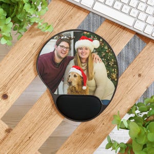 Peut inclure: Un tapis de souris ergonomique noir avec repose-poignet. Le tapis présente une photo circulaire d'un couple souriant et d'un chien, tous portant des chapeaux de Père Noël. L'arrière-plan comprend un sapin de Noël.