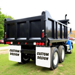 May include: A black dump truck with a blue cab parked on grass. The truck has white mud flaps with the words "CUSTOM DESIGN" printed on them. The truck has red and white lights.