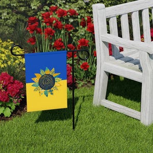 May include: A garden flag with a yellow and blue background featuring a stylised sunflower with green and blue accents. The flag is attached to a black metal garden flag stand. The flag is displayed in a garden setting with a white wooden bench and red flowers in the background.