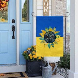 May include: A blue and yellow garden flag with a stylized sunflower design. The sunflower is made up of different colored shapes and patterns.