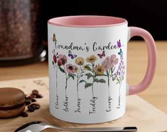 May include: A white ceramic mug with a pink rim and handle. The mug features a floral design with butterflies and the text "Grandma's Garden".  The names Oliver, Arthur, James, Freddy, George, and Emma are written vertically along the stems of the flowers.