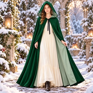 May include: A floor-length dark green hooded cloak with a light green lining and a black ribbon tie at the neck, worn over a long cream-colored dress with a ruffled hem. The cloak features a deep hood and a flowing, full-length silhouette.