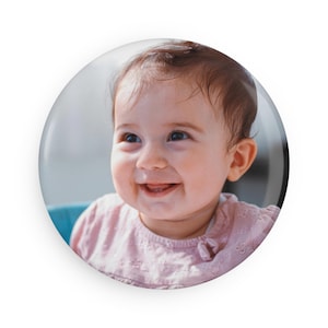 Könnte beinhalten: Ein runder Button mit einem lächelnden Baby mit braunen Haaren und blauen Augen. Das Baby trägt ein rosa Hemd und ist vor einem verschwommenen Hintergrund platziert. Der Button hat ein glänzendes Finish.