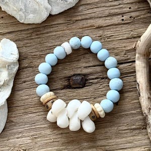 May include: A bracelet with light blue, round beads, white and tan decorative elements, and white shell-shaped beads. The bracelet is displayed on a rustic wooden surface. The beads are likely around 0.8 cm in diameter.