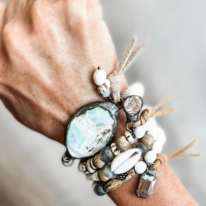 May include: A multi-strand bracelet featuring a large, oval, blue and white marbled stone set in dark metal, accented with white beads, tan leather cords, a white cowrie shell, and a rectangular faceted glass charm on a dark metal base.