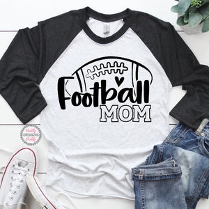 May include: A white and black long-sleeved shirt with a football graphic and the text "Football Mom". The shirt has a black raglan sleeve design.