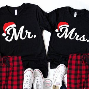 May include: A pair of black t-shirts with red Santa hats and the words "Mr." and "Mrs." printed on them. The shirts are paired with red and black checked pyjama bottoms.