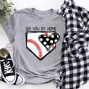 Puede incluir: Camiseta gris con un gr&aacute;fico de b&eacute;isbol y el texto "See you at home". El gr&aacute;fico presenta una pelota de b&eacute;isbol en un plato de home con un coraz&oacute;n de lunares negros y blancos.
