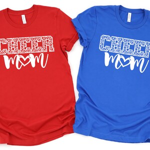 May include: Two t-shirts with the text "CHEER MOM" in white with a white heart in the middle of the word "MOM". The word "CHEER" is covered in a white leopard print. One shirt is red and the other is blue.