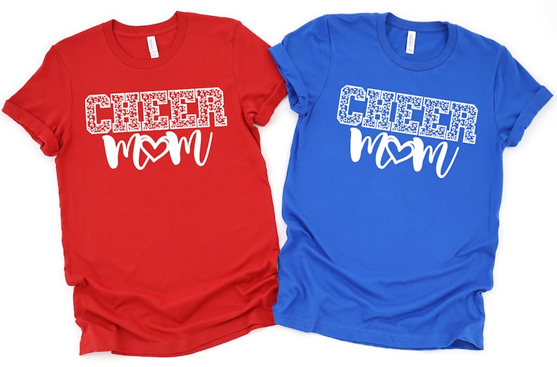 May include: Two t-shirts with the text "CHEER MOM" in white with a white heart in the middle of the word "MOM". The word "CHEER" is covered in a white leopard print. One shirt is red and the other is blue.