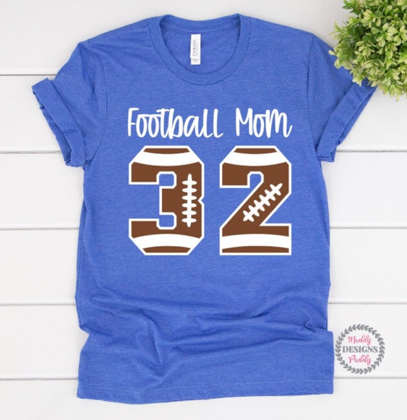 K&ouml;nnte beinhalten: Blaues T-Shirt mit dem Text "Football Mom 32" in Wei&szlig;. Die Zahl 32 besteht aus zwei braunen Fu&szlig;b&auml;llen.