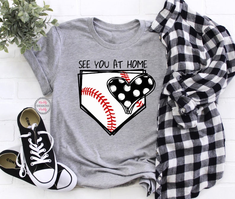Puede incluir: Camiseta gris con un gr&aacute;fico de b&eacute;isbol y el texto "See you at home". El gr&aacute;fico presenta una pelota de b&eacute;isbol en un plato de home con un coraz&oacute;n de lunares negros y blancos.