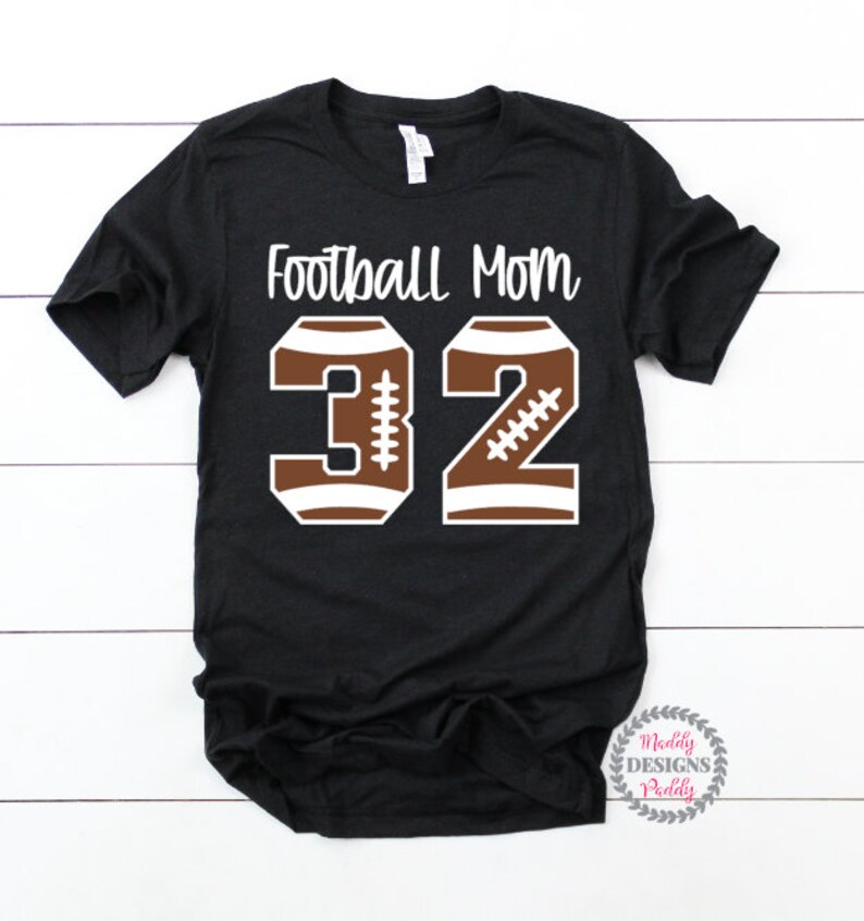 K&ouml;nnte beinhalten: Schwarzes T-Shirt mit dem Text "Football Mom" und der Nummer "32" in Braun und Wei&szlig;, mit einem Fu&szlig;ball-Design.