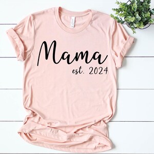 以下が含まれることがあります： 黒い文字で「ママ est. 2024」と書かれた薄いピンクのTシャツ