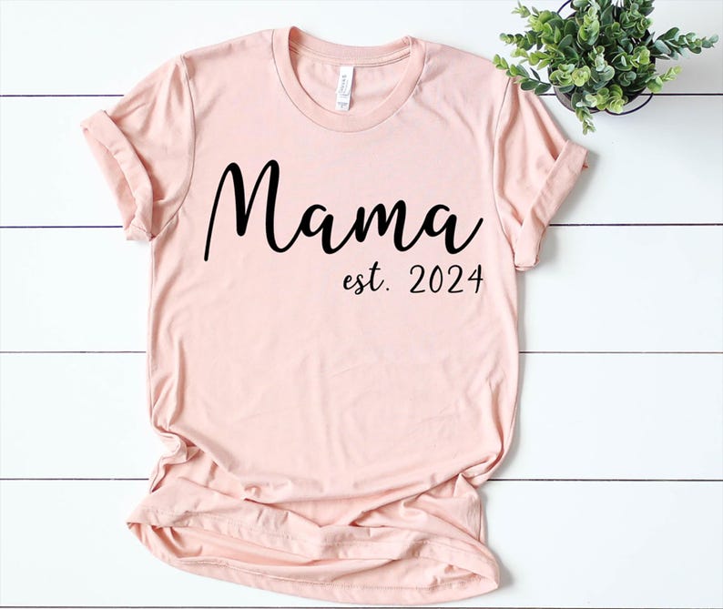 以下が含まれることがあります： 黒い文字で「ママ est. 2024」と書かれた薄いピンクのTシャツ