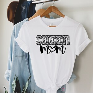May include: White t-shirt with black text that says "CHEER MOM" with a heart in the middle of the word "MOM". The word "CHEER" is in a leopard print pattern.