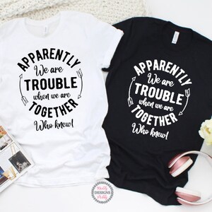 May include: Two t-shirts with the text "Apparently we are trouble when we are together. Who knew!" on a white background with black text and arrows. The t-shirts are on a white surface with a book and flowers.