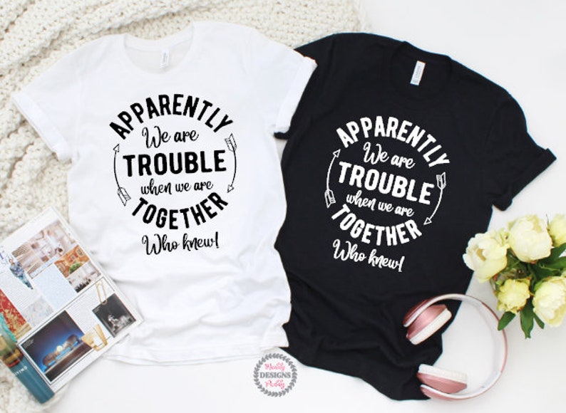 May include: Two t-shirts with the text "Apparently we are trouble when we are together. Who knew!" on a white background with black text and arrows. The t-shirts are on a white surface with a book and flowers.