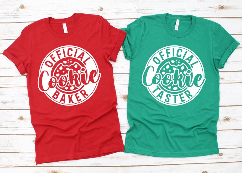 Pode incluir: Duas camisetas de manga curta, uma vermelha e outra verde, com desenhos circulares brancos. A camiseta vermelha diz "Official Cookie Baker" e a camiseta verde diz "Official Cookie Taster". Ambas as camisetas est&atilde;o em uma superf&iacute;cie de madeira clara.