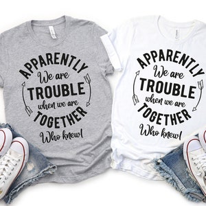 May include: Two gray and white t-shirts with the text "Apparently We are Trouble When We are Together Who Knew!" in a circle with arrows on either side. The shirts are laid out on a white background with denim shorts and white sneakers.