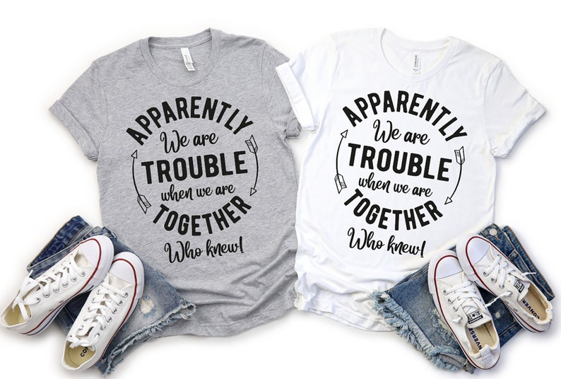 May include: Two gray and white t-shirts with the text "Apparently We are Trouble When We are Together Who Knew!" in a circle with arrows on either side. The shirts are laid out on a white background with denim shorts and white sneakers.