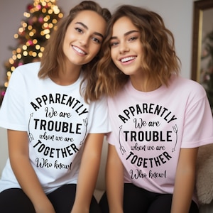 May include: Two women wearing matching t-shirts with the text "Apparently we are trouble when we are together. Who knew!" One shirt is white and the other is pink.
