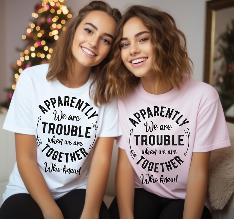 May include: Two women wearing matching t-shirts with the text "Apparently we are trouble when we are together. Who knew!" One shirt is white and the other is pink.