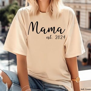以下が含まれることがあります： クリーム色のソフトなTシャツに、黒い文字で「Mama est. 2024」と書いてあります。
