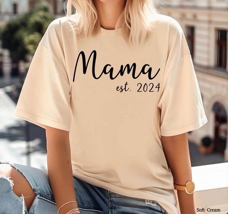 以下が含まれることがあります： クリーム色のソフトなTシャツに、黒い文字で「Mama est. 2024」と書いてあります。