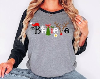 May include: A gray and black long-sleeved shirt with a Christmas-themed design that says "Believe" with a Santa hat, snowflake, Christmas tree, bow, holly, reindeer antlers, star, and ornament.