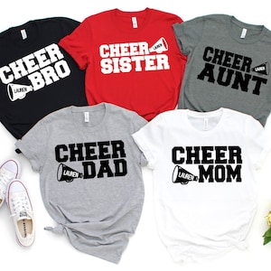 May include: Five t-shirts with different designs featuring the word "Cheer" and a megaphone. The shirts are in black, red, grey, and white. The text on the shirts reads "Cheer Bro", "Cheer Sister", "Cheer Aunt", "Cheer Dad", and "Cheer Mom".