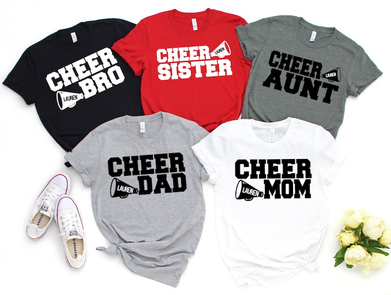 May include: Five t-shirts with different designs featuring the word "Cheer" and a megaphone. The shirts are in black, red, grey, and white. The text on the shirts reads "Cheer Bro", "Cheer Sister", "Cheer Aunt", "Cheer Dad", and "Cheer Mom".