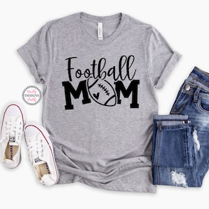 K&ouml;nnte beinhalten: Ein graues T-Shirt mit dem Text "Football Mom" in schwarzen Buchstaben. Ein Fu&szlig;ball mit einem Herzen ist im Design enthalten. Das Shirt wird mit einer blauen Jeans und wei&szlig;en Turnschuhen gezeigt.