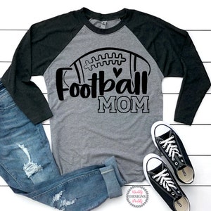 May include: A gray and black long-sleeved shirt with a football graphic and the text "Football Mom". The shirt is on a white wooden surface with a pair of black sneakers and blue jeans.