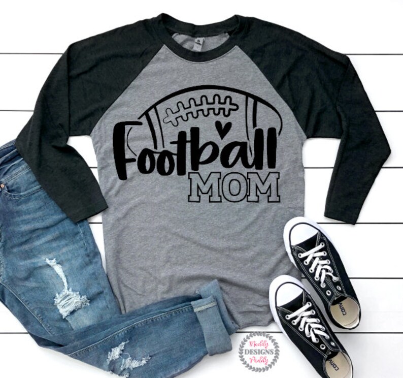 May include: A gray and black long-sleeved shirt with a football graphic and the text "Football Mom". The shirt is on a white wooden surface with a pair of black sneakers and blue jeans.