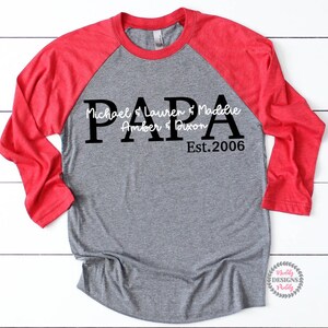 May include: A gray and red baseball tee shirt with the word "PAPA" in large black letters. The text "Michael & Lauren & Maddie Amber & Dixon Est. 2006" is written in white cursive letters below the word "PAPA".