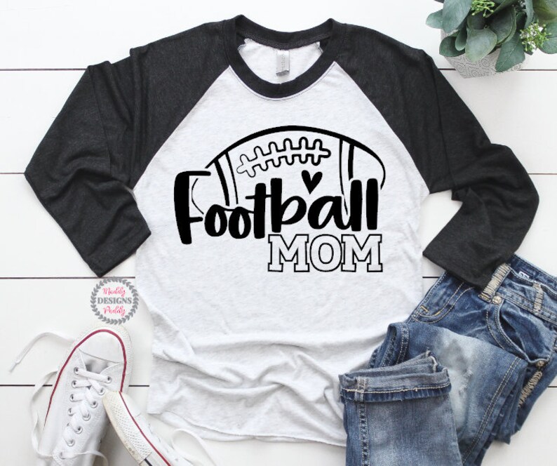 May include: A white and black long-sleeved shirt with a football graphic and the text "Football Mom". The shirt has a black raglan sleeve design.