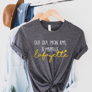 May include: A dark gray t-shirt with the text "Oui oui, mon ami, je m'appelle Lafayette" in white with a yellow star. The text is in a cursive font.