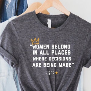 May include: A dark heather grey t-shirt with a white quote that reads "Women belong in all places where decisions are being made" with a crown and a star above the quote and "RBG" below the quote. The quote is in orange.